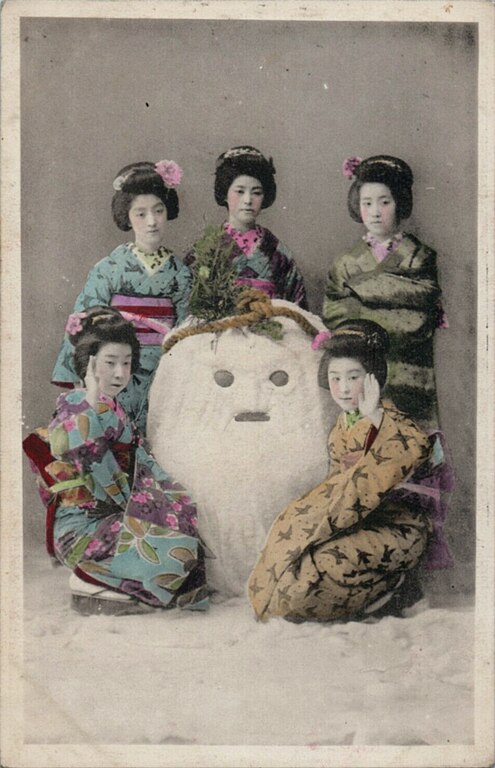 Kolorierte Fotografie: Fünf Geishas posieren um eine eine Schneefigur, der mit einem Tau Blumen auf die Stirn gebunden sind. Die Schneefigur schaut aus großen, dunklen Augen ausdruckslos in die Kamera.