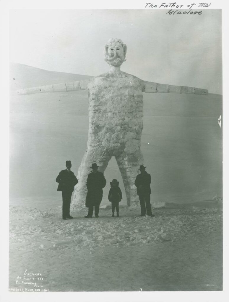 Silhouettes of three men in hats and a child in snow-filled landscape standing in front of a monumental snowman sculpture. The snowman has a head with eyes, nose and mouth, modeled hair and mustache, two arms and legs, and an oval body. The snowman is approximately four times the size of a man.