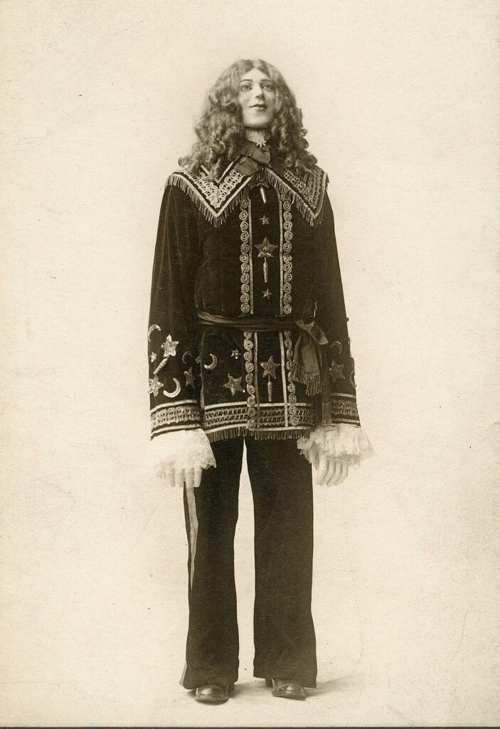 early 20th century photo of a long-haired, curly person with arms that seem a bit too long, artificial hands, wearing an old-fashioned costume adorned with stars and crescents, and ruffled sleeves. The person shows a mask-like smile stairing slightly upwards