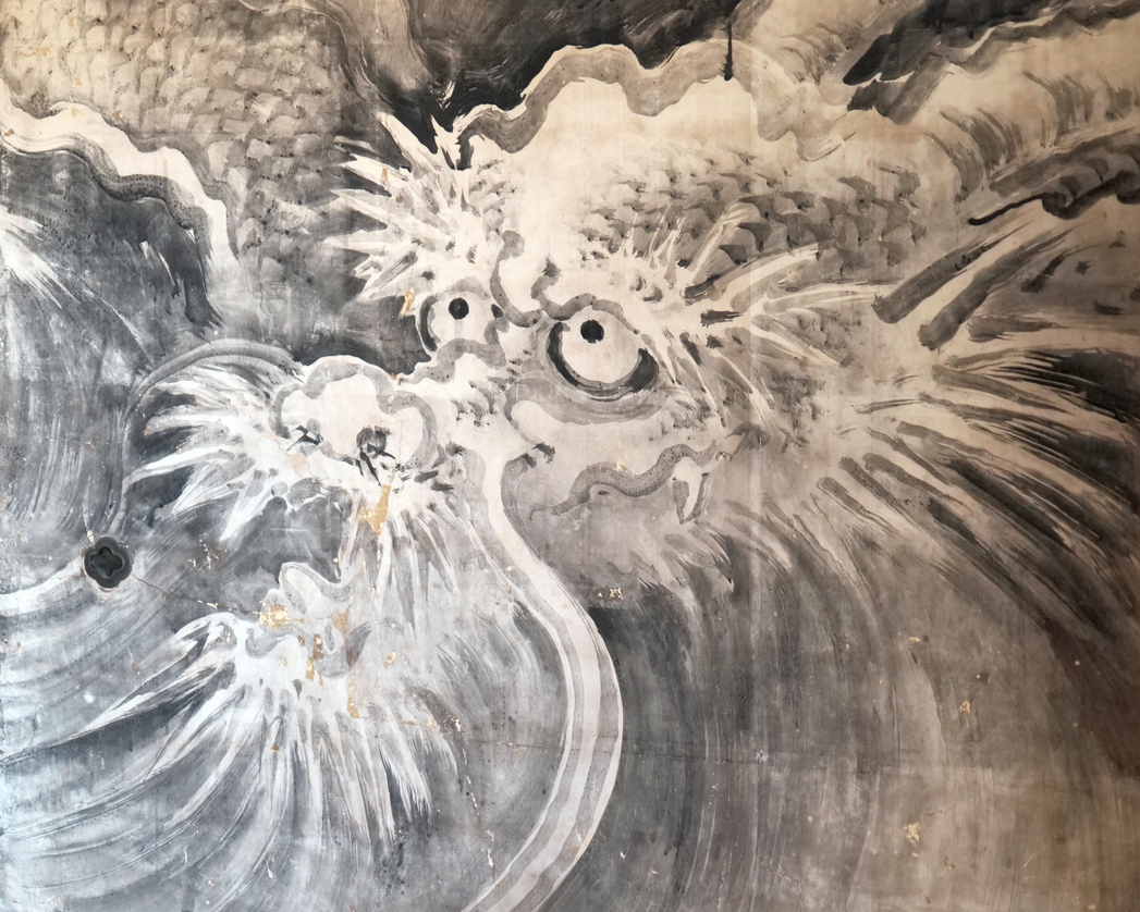 Foto eines gemalten "östlichen" Drachens auf einer Tempelwand mit buschigen Augenbrauen und großen kugeligen Augen.